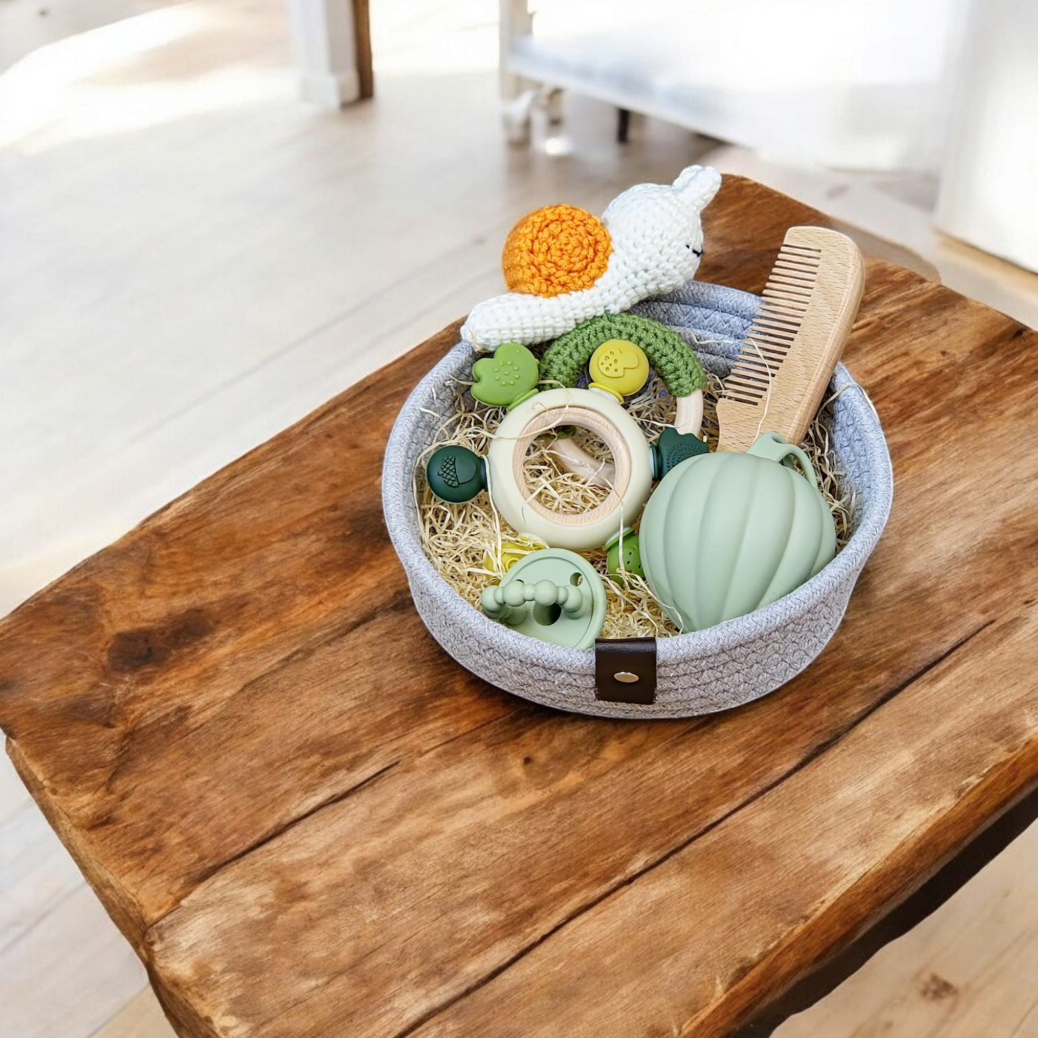 Der Hello World Geschenk Konfigurator besteht aus einem grauen Stoffkorb mit Babyspielzeug - Holzring, Beißringe, grüne und gelbe Rasseln und ein gehäkeltes Häschen - und eignet sich ideal als Geschenkset für Babys auf einem rustikalen Holztisch in einem hellen Raum.