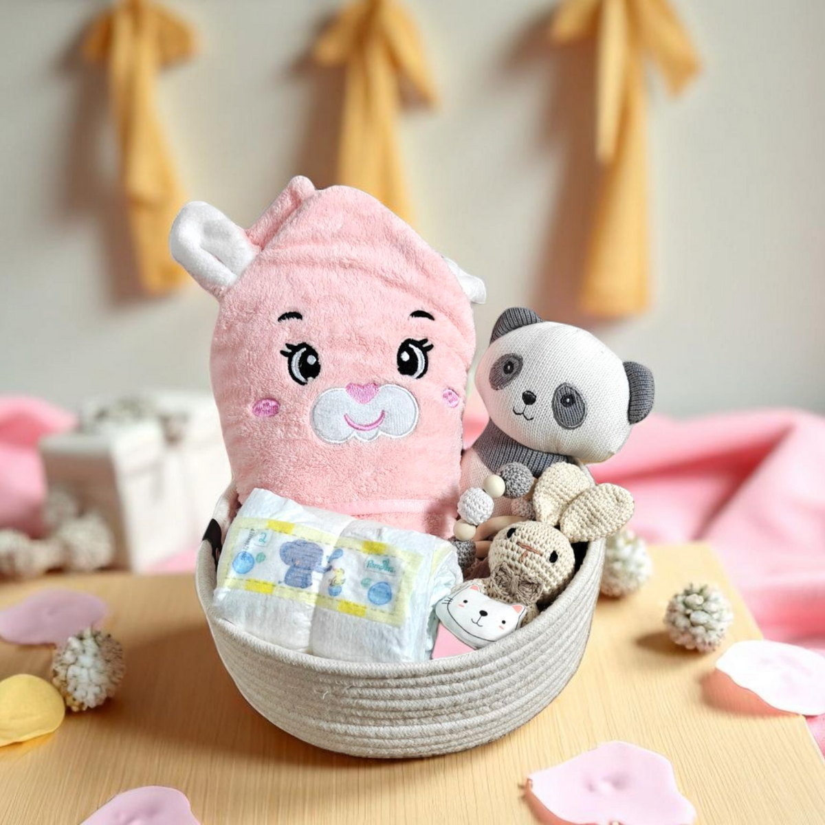 Das Geschenkset "Geschenk Konfigurator" von Hello World enthält ein pinkes Tier-Kapuzenhandtuch, einen Kuschel-Panda, Pflegeartikel und Windeln auf einem Holzbrett. Rosa Stoff, weiße Blumen und Geschenkboxen dekorieren den Hintergrund.