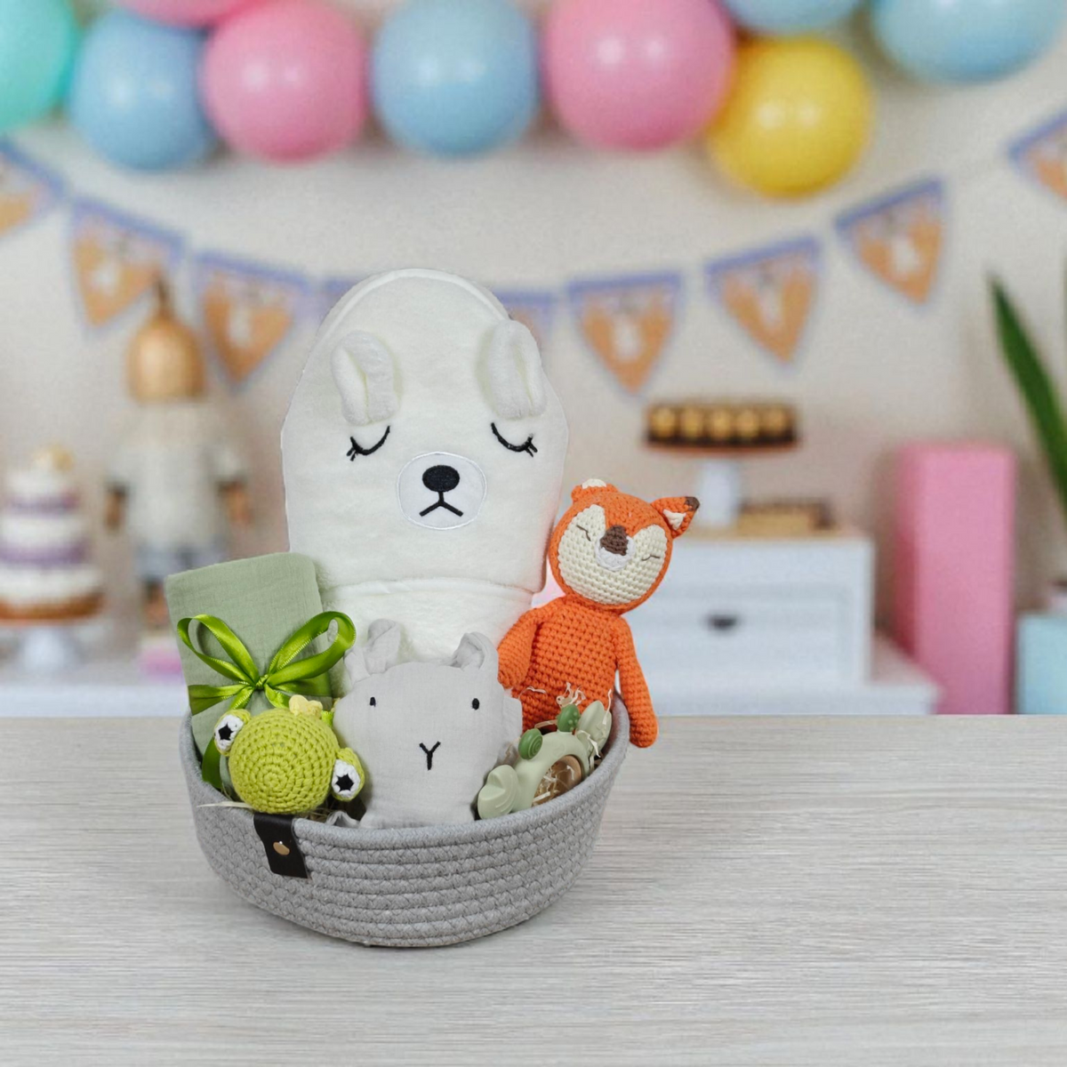 Das Hello World Geschenk Konfigurator-Set mit plüschigen und gehäkelten Tieren, grünem Tuch und Babyartikeln steht auf einem Holztisch. Im unscharfen Hintergrund sind bunte Ballons und eine Wimpelkette zu sehen.
