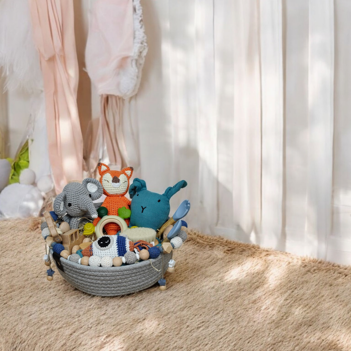 Ein grauer geflochtener Korb vom Geschenk Konfigurator von Hello World, gefüllt mit farbenfrohen, personalisierten Baby-Geschenksets - gehäkelte Tiere wie ein Fuchs, ein Elefant und ein blaues Wesen - liegt auf einem beigen Teppich in einem gemütlichen Zimmer mit weißen Vorhängen.