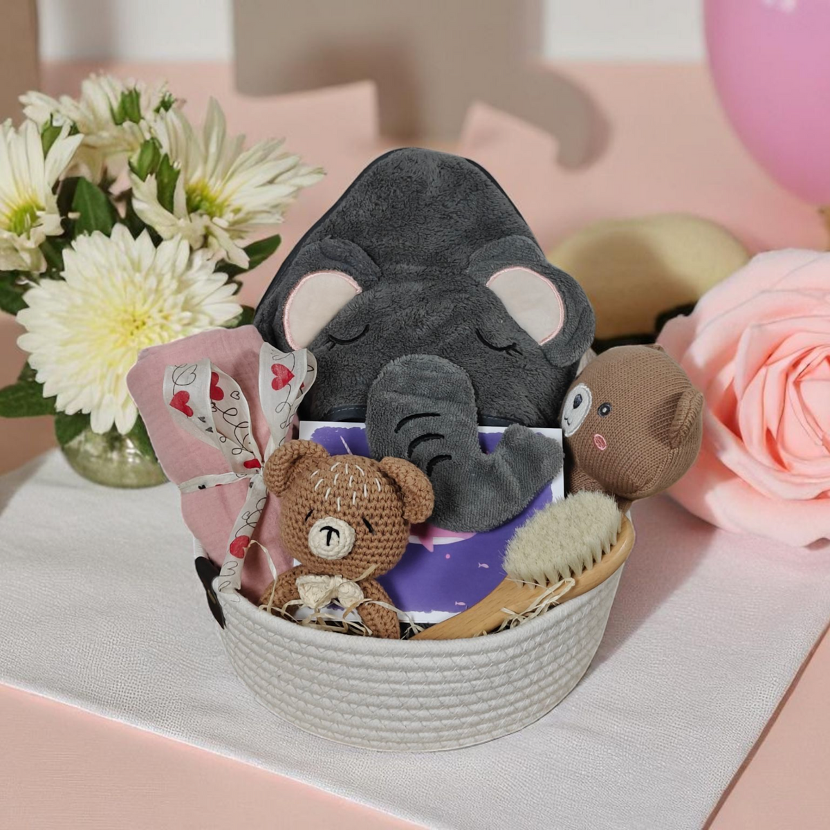 Das Geschenk Konfigurator von Hello World ist ein liebevolles Babyset mit Plüsch-Elefant, Teddy, gehäkeltem Tier, Babybürste und geblümten Accessoires - stilvoll inszeniert zwischen rosa Rosen und weißen Blüten.