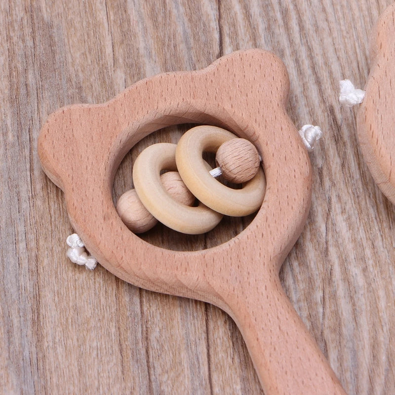 Eine Hello World Griff aus Holz [8x12cm] Babyrassel in Form eines Bärenkopfes mit zwei hellen Holzringen und inneren Perlen, mit einem ergonomischen Holzgriff und auf einer Holzfläche ruhend.
