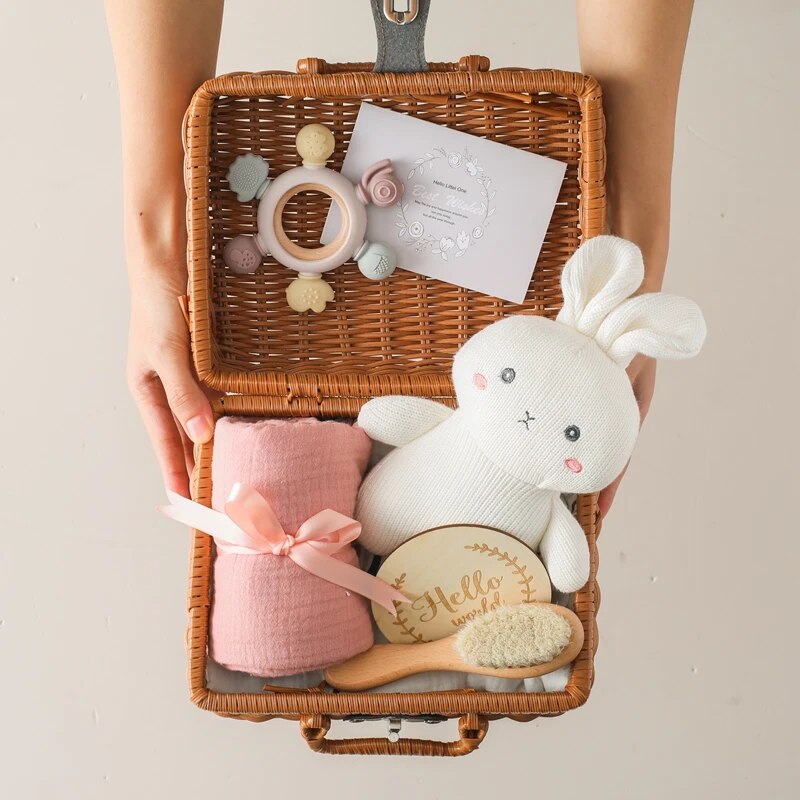 Ein Paar Hände hält das Hello World Hanna Baby Geschenk zur Geburt: ein offener Weidenkorb mit einer rosa Decke, Plüschhase aus Baumwolle, Holzhaarbürste, Holz-Silikon-Beißring und einer Karte mit Pastellfarben - perfekt für Neugeborene.