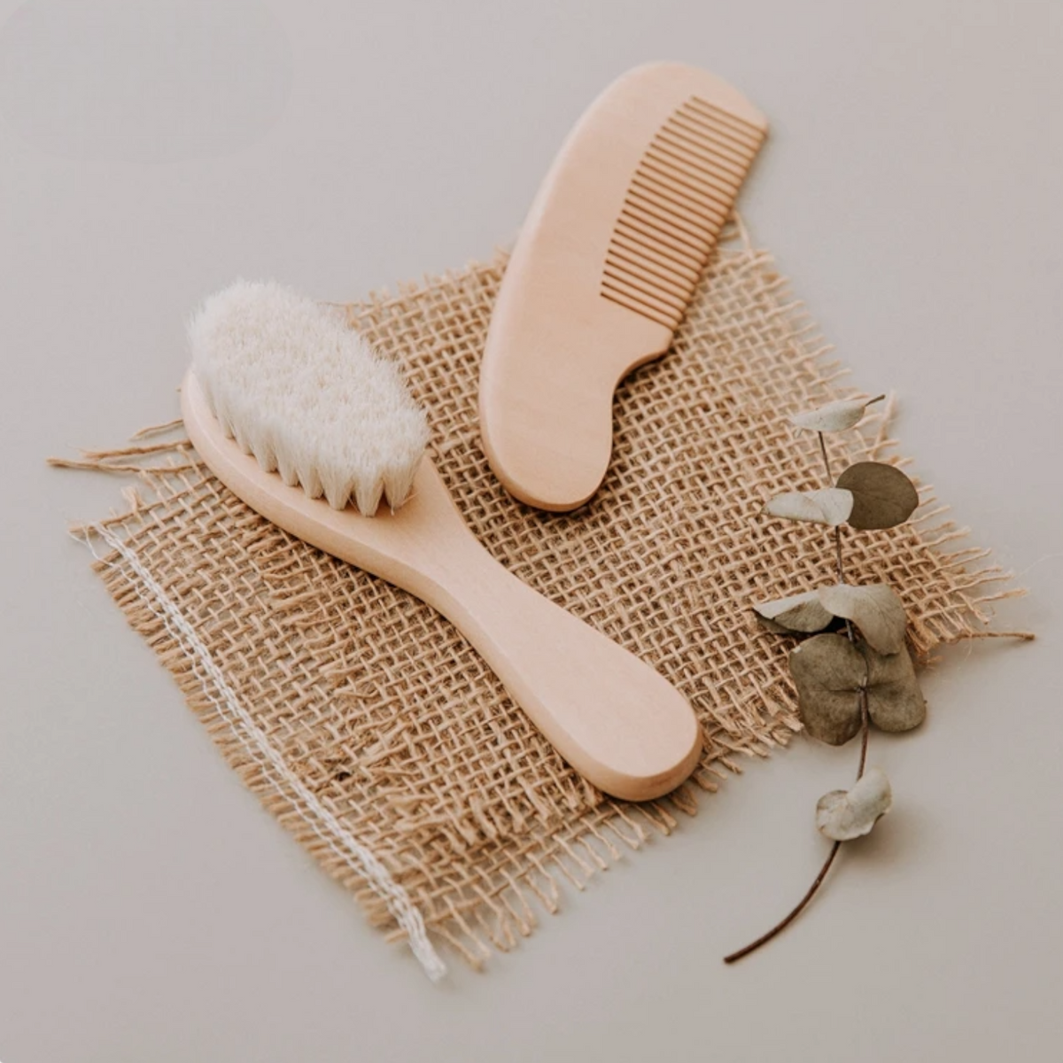 Ein Hello World Kamm [4,5 x 13,5 cm] und eine hölzerne Babyhaarbürste mit weichen weißen Borsten ruhen auf Sackleinen neben Eukalyptusblättern und bieten sanfte Pflege für feines Babyhaar in einer ruhigen, neutralen Umgebung.