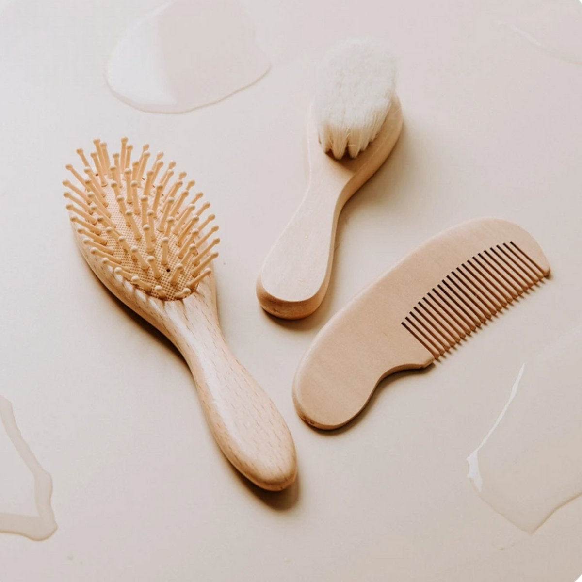 Drei Hello World-Haarpflegewerkzeuge - ein Babykamm [4,5 x 13,5 cm], eine Bürste mit weichen Borsten und ein Kamm - sind auf einer hellen Oberfläche mit Wassertropfen angeordnet und unterstreichen die sanfte Pflege für feines Babyhaar.