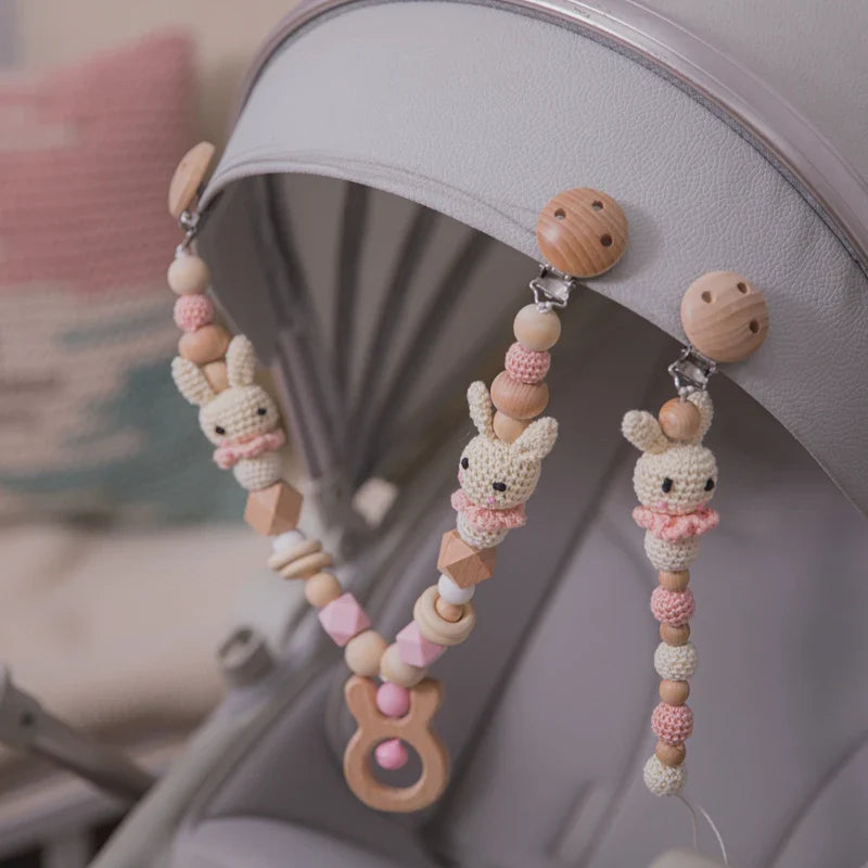 Ein Kinderwagen mit der Hello World Kinderwagenkette Hase [20x29cm] in rosa und beige, mit Hasenformen, Holzperlen und praktischen Clips - ein bezauberndes Babyspielzeug für unterwegs.