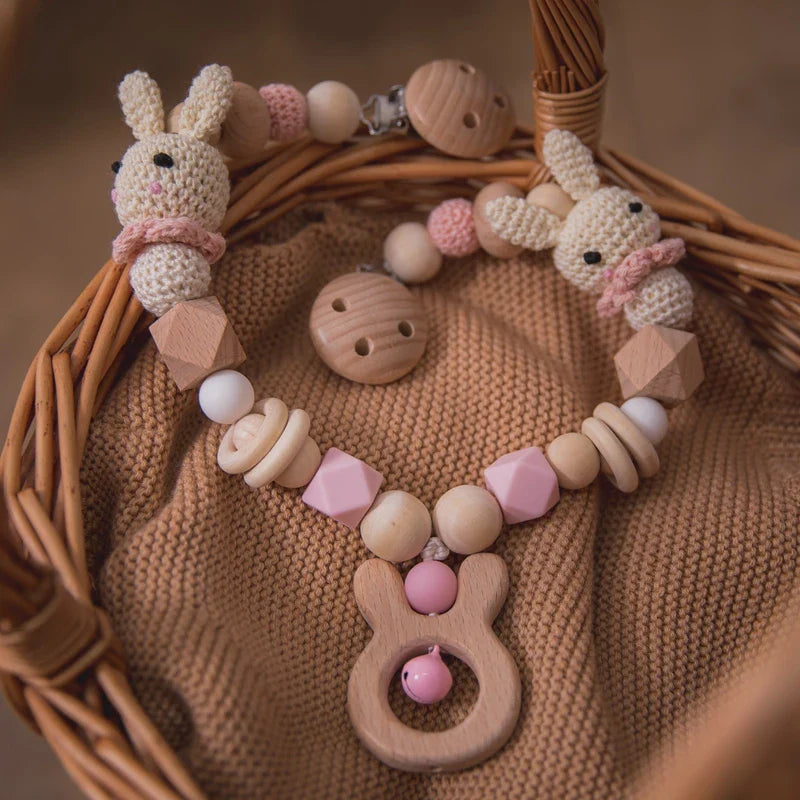Die Hello World Kinderwagenkette Hase [20x29cm], ein hölzernes und gehäkeltes Babyspielzeug in Hasenform mit rosa und weißen Perlen, Holzringen und zwei gehäkelten Hasengesichtern, wird in einem Weidenkorb auf einer weichen braunen Decke gezeigt.