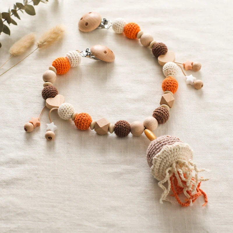 Die Hello World Kinderwagenkette Qualle [30x34cm] ist eine handgefertigte Kinderwagenkette mit Holz- und Häkelperlen in Weiß, Braun und Orange, Quallen und Sternen - ideal für eine Unterwasserwelt Baby - auf einem hellen Stoffhintergrund.