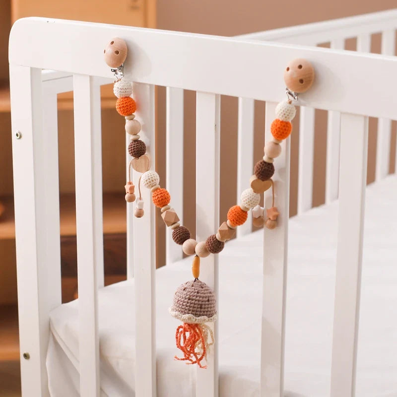 Eine weiße Krippe zeigt die Hello World Kinderwagenkette Qualle [30x34cm], eine hängende Perlengirlande mit orangefarbenen, braunen und beigen Häkel- und Holzperlen sowie eine kleine Häkelglocke mit orangefarbenen Quasten als beruhigende Dekoration.