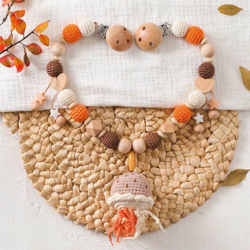 Die Hello World Kinderwagenkette Qualle [30x34cm] zeigt Holz- und Häkelperlen in Orange, Creme und Braun mit Herz-, Stern- und Quallenformen, die auf einer gewebten Matte mit Herbstblättern abgebildet sind.