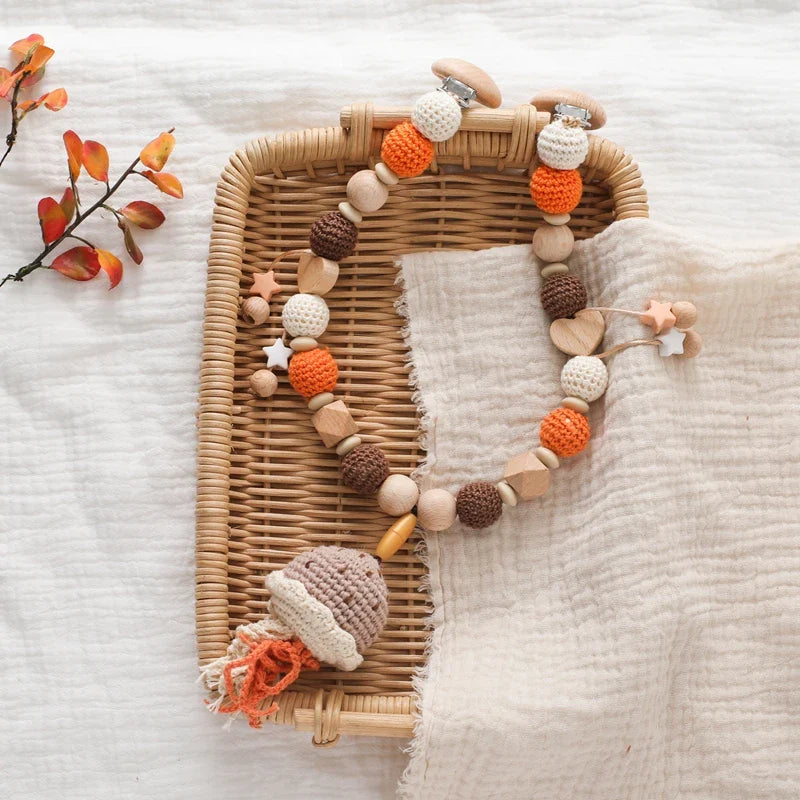 Ein gewebtes Tablett enthält die Hello World Kinderwagenkette Qualle [30x34cm] mit gehäkelten und hölzernen Perlen in Orange, Braun und Creme sowie einer hölzernen, gehäkelten Qualle. Ein beiger Stoff und Herbstblätter schaffen eine gemütliche Unterwasser-Baby-Szene.