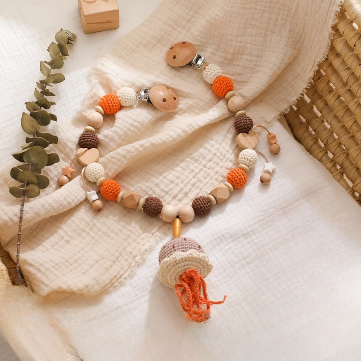 Die Kinderwagenkette Qualle [30x34cm] von Hello World besticht durch beruhigende Erdtöne und charmante Häkeldetails, die auf einem cremefarbenen Strukturstoff neben getrocknetem Eukalyptus präsentiert werden - das perfekte beruhigende Babyspielzeug.