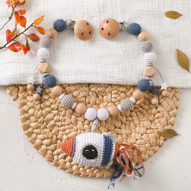 Die Hello World Kinderwagenkette Rakete [30x38cm] besteht aus handgefertigten Holzperlen und gehäkelten Perlen in neutralen Farbtönen sowie einem Raketenschiff - ideal als Baby Weltraum Spielzeug oder Kinderwagen Anhänger. Präsentiert auf einer gewebten Matte mit Herbstblättern und weißem Stoff.