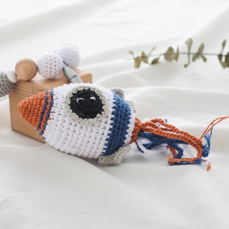 Die Hello World Kinderwagenkette Rakete (30x38cm) ist eine gehäkelte Spielzeugrakete in weißem, orangefarbenem, blauem und grauem Garn mit einem großen schwarzen Auge und Quastenschwanz, ideal als Baby Weltraum Spielzeug oder Kinderwagen Anhänger.