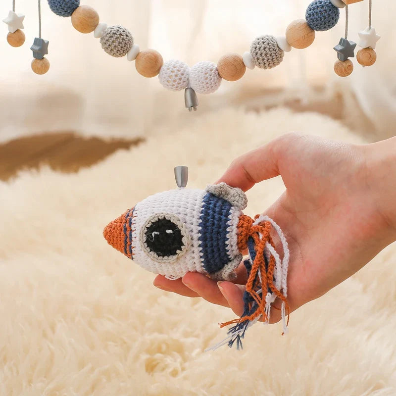 Eine Hand hält eine kleine gehäkelte Rakete als Kinderwagen-Anhänger, während die Hello World Kinderwagenkette Rakete [30x38cm], eine Perlengirlande in den Farben Blau, Weiß, Orange und Grau, über einem cremefarbenen Flauschteppich hängt.