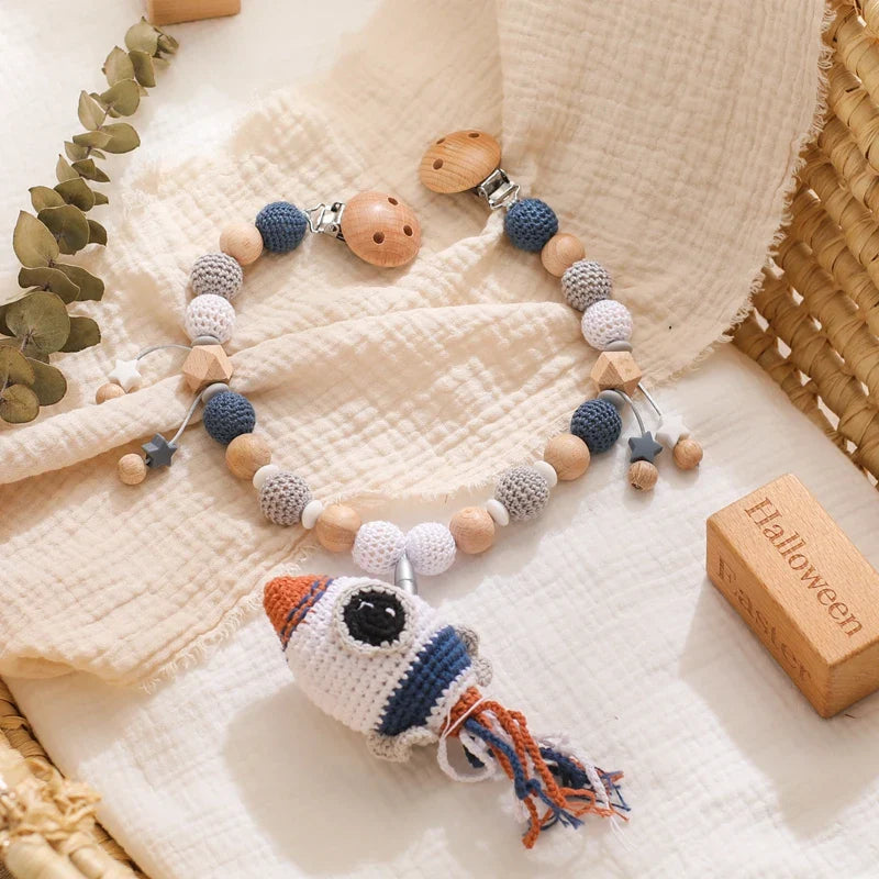 Die Hello World Kinderwagenkette Rakete [30x38cm] besteht aus hölzernen und gehäkelten Perlen in Blau, Weiß und Grau mit einem Raketen-Anhänger. Abgebildet auf einer cremefarbenen Decke neben einem Weidenkorb und einem hölzernen Halloween-Block.