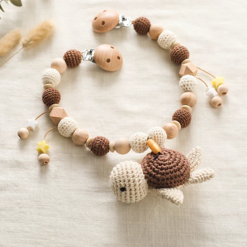 Die Hello World Kinderwagenkette Schildkröte [30x31cm] hat beige und braune Holz- und Häkelperlen, eine süße gehäkelte Schildkröte in der Mitte und sternförmige Anhänger für entspannte Spaziergänge.