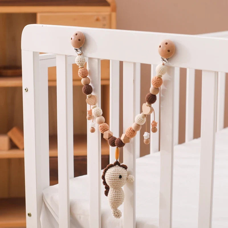 Ein weißes Kinderbett ist mit der Hello World Kinderwagenkette Seepferdchen (30x35cm) dekoriert, einer gehäkelten Perlengirlande in neutralen Farbtönen, inspiriert von Unterwasserwelt Baby, mit einem Seepferdchen als Mittelstück. Im Hintergrund sind Holzmöbel zu sehen.