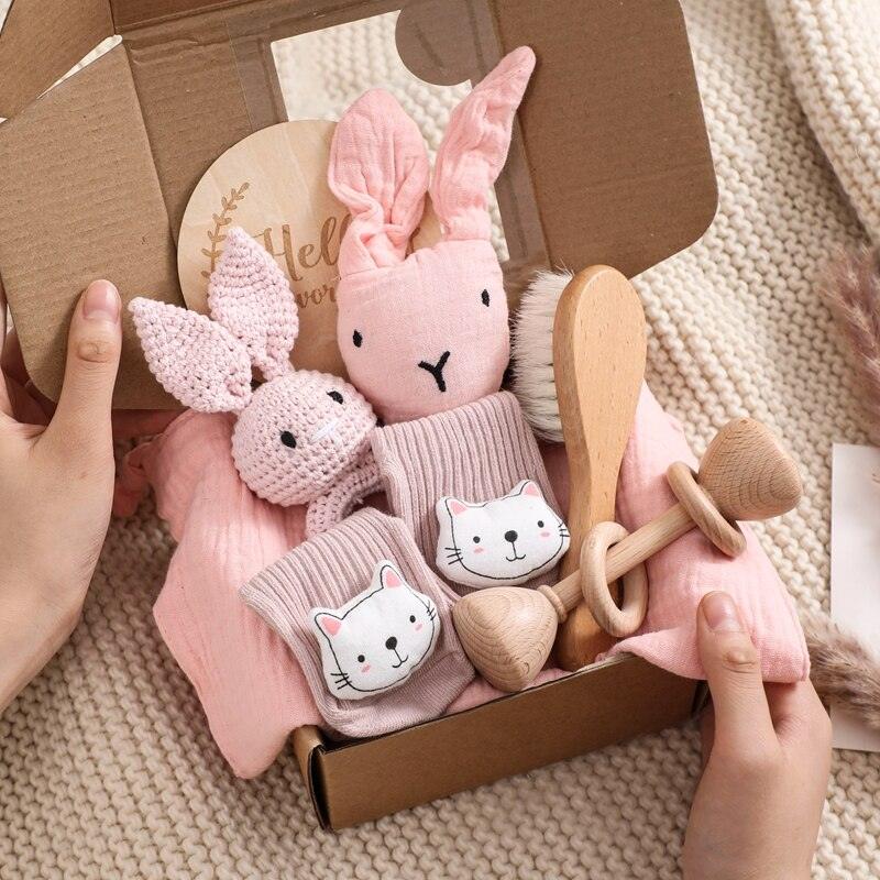 Das Kira Baby Geschenk zur Geburt von Hello World enthält pinke Rasselhase-Puppe, Cat-Face-Socken, Holzherz, Holzrassel, Bürste und rosa Tuch - liebevoll arrangiert in einer Geschenkbox. Zwei Hände halten das Set auf beigem Untergrund.