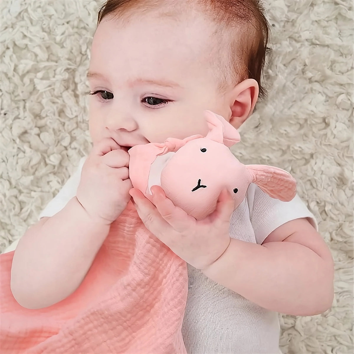 Ein Baby liegt auf einem weichen cremefarbenen Teppich, trägt einen weißen Strampler und kaut sanft auf dem Stoff der rosafarbenen Kira Baby Geschenk zur Geburt Bettdecke von Hello World - eine bezaubernde Szene.