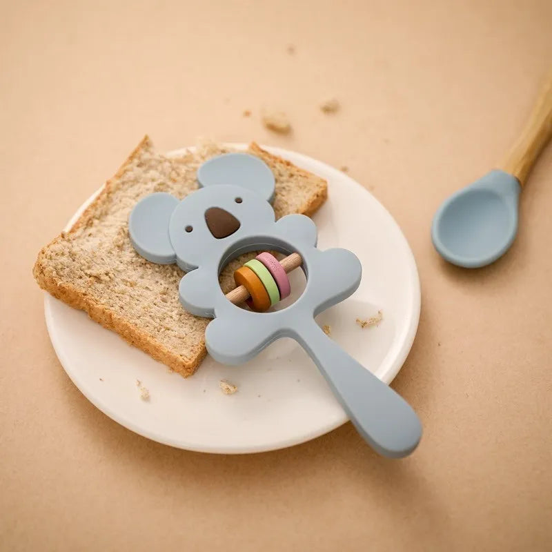 Eine Scheibe Brot auf einem weißen Teller mit einem Hello World Koala Blau aus Silikon [7x15cm] und einem blauen Löffel mit Holzgriff auf einer beigen Fläche.