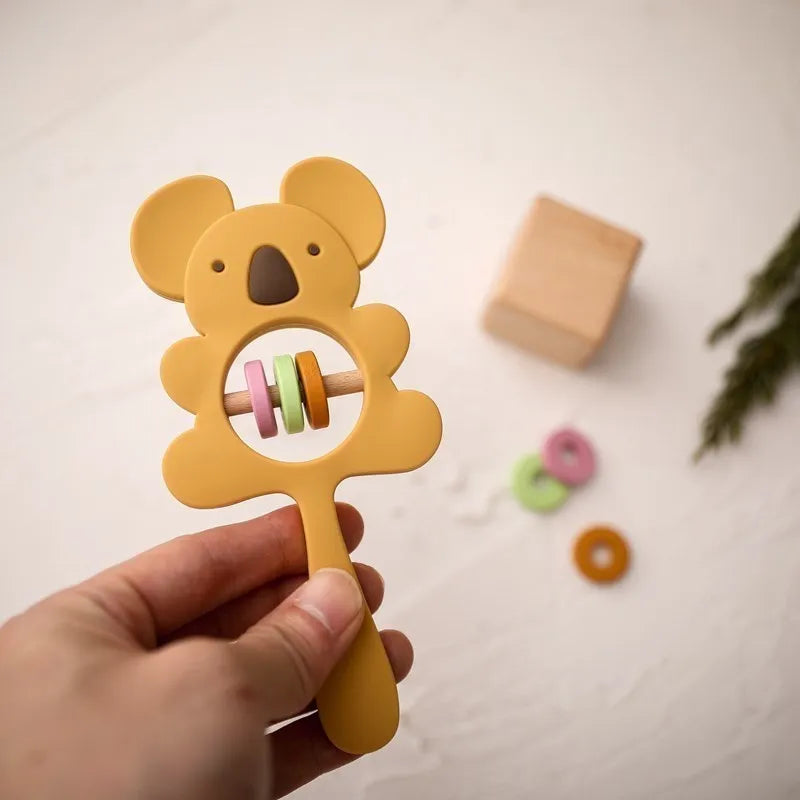 Eine Hand hält die Hello World Koala Gelb aus Silikon [7x15cm] Babyrassel mit pastellfarbenen Ringen im Inneren. Im Hintergrund sind ein Holzblock, bunte Ringe und ein grüner Zweig auf einer hellen Fläche zu sehen.