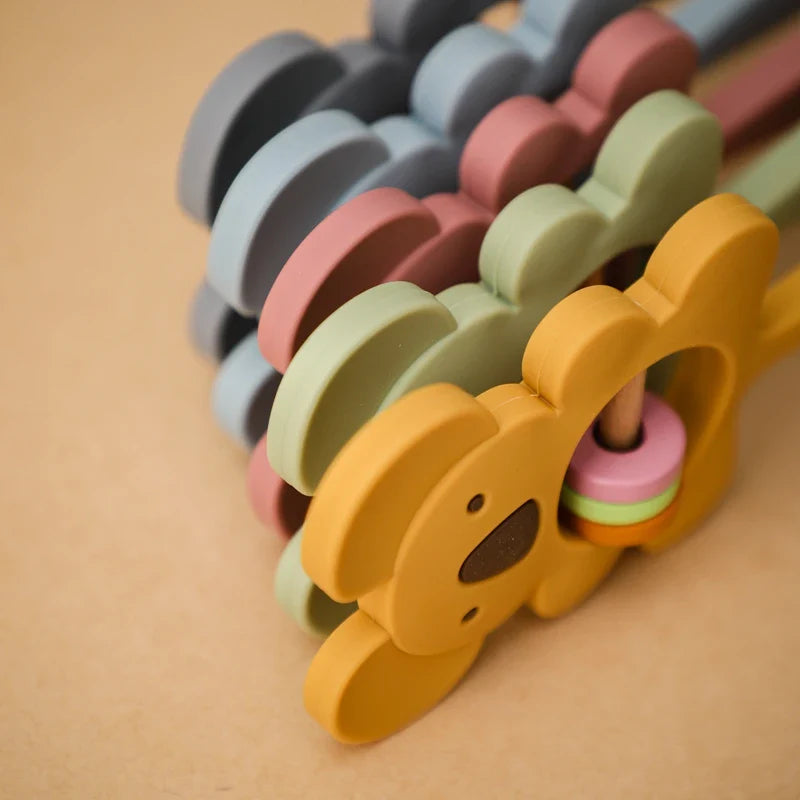 Eine Nahaufnahme des Hello World Koala Gelb aus Silikon [7x15cm] und anderer farbenfroher Babyrasseln, die alle aus lebensmittelechtem Silikon hergestellt sind, gestapelt auf einer hellbeigen Oberfläche.