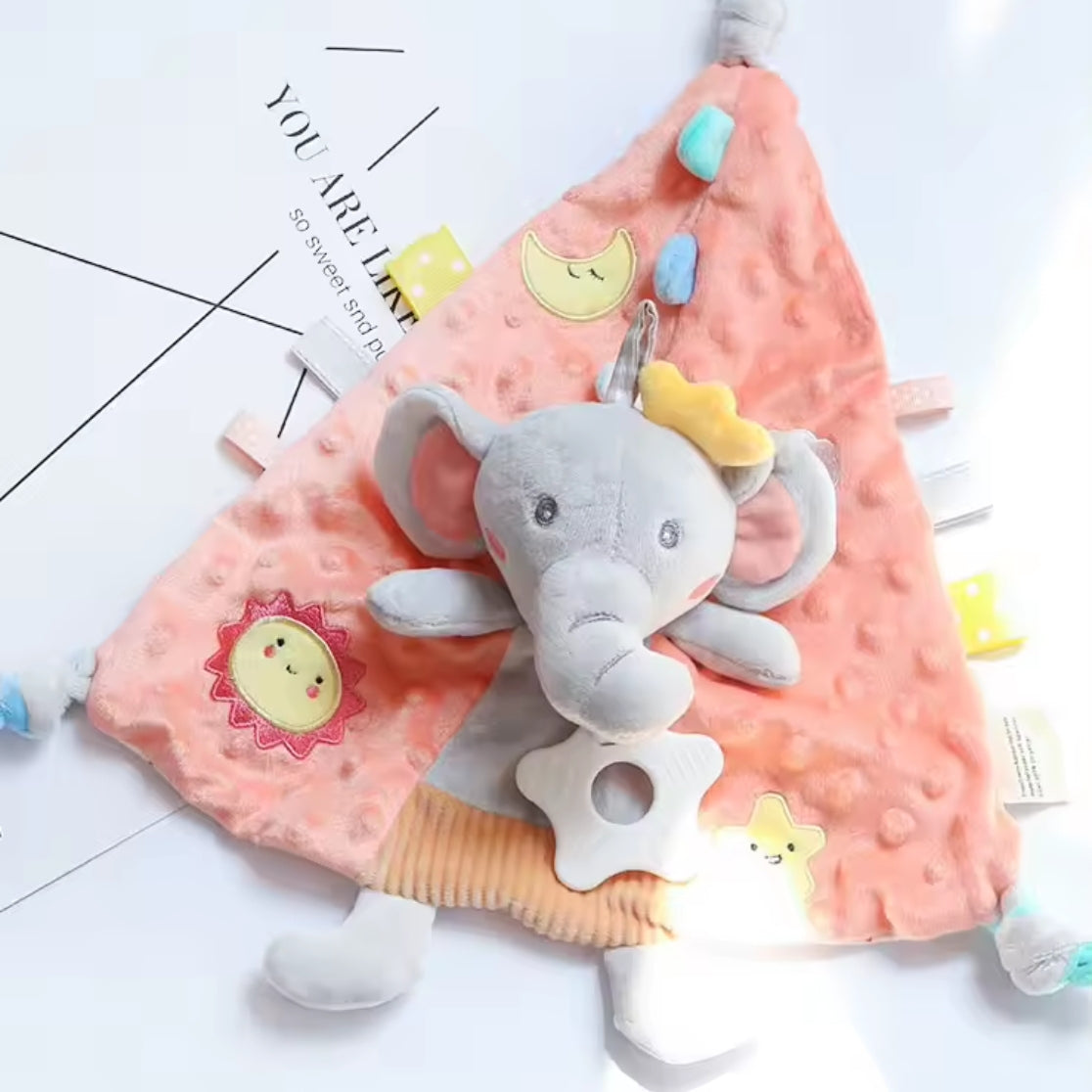 Das Kuscheldreieck Elefant [30x30cm] von Hello World ist ein weiches, pfirsichfarbenes Schmusetuch mit einem grauen Plüschelefanten in der Mitte, Sonnen-, Mond- und Sternenflecken und strukturierten Anhängern - ideal als Baby-Einschlafhilfe.