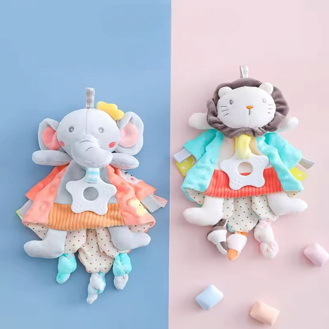 Zwei Plüschbabys ruhen auf einem blau-rosa Hintergrund. Das eine ist das Hello World Kuscheldreieck Elefant (30x30cm) mit großen Ohren und gelbem Hut, das andere ist ein Löwe. Beide tragen bunte Kleidung und halten sternförmige Beißringe, ideal als Einschlafhilfe.