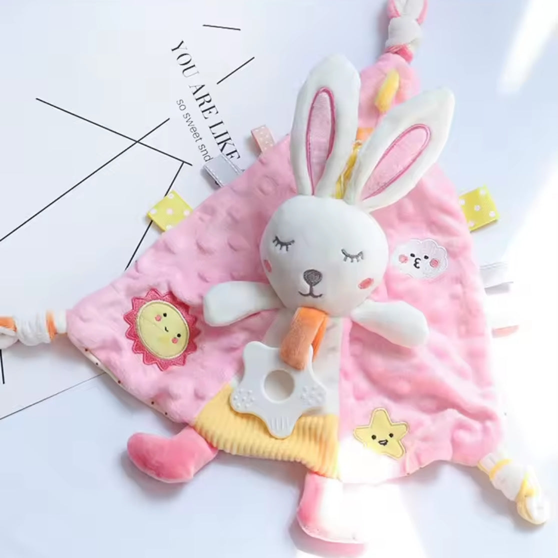 Ein weicher Kuscheldreieck Hase [30x30cm] von Hello World, in rosa und gelb mit einem Plüschhasen in der Mitte, Tierflecken und strukturierten Anhängern, liegt auf einer weißen Fläche neben einem teilweise sichtbaren bedruckten Papier.