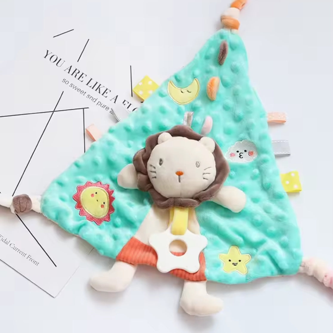 Eine weiche, tealfarbene Kuscheldreieck Löwe [30x30cm] Babydecke von Hello World mit bunten Anhängern und einem angehängten Plüschlöwen ruht auf weißen Papierbögen mit aufgedrucktem Text.