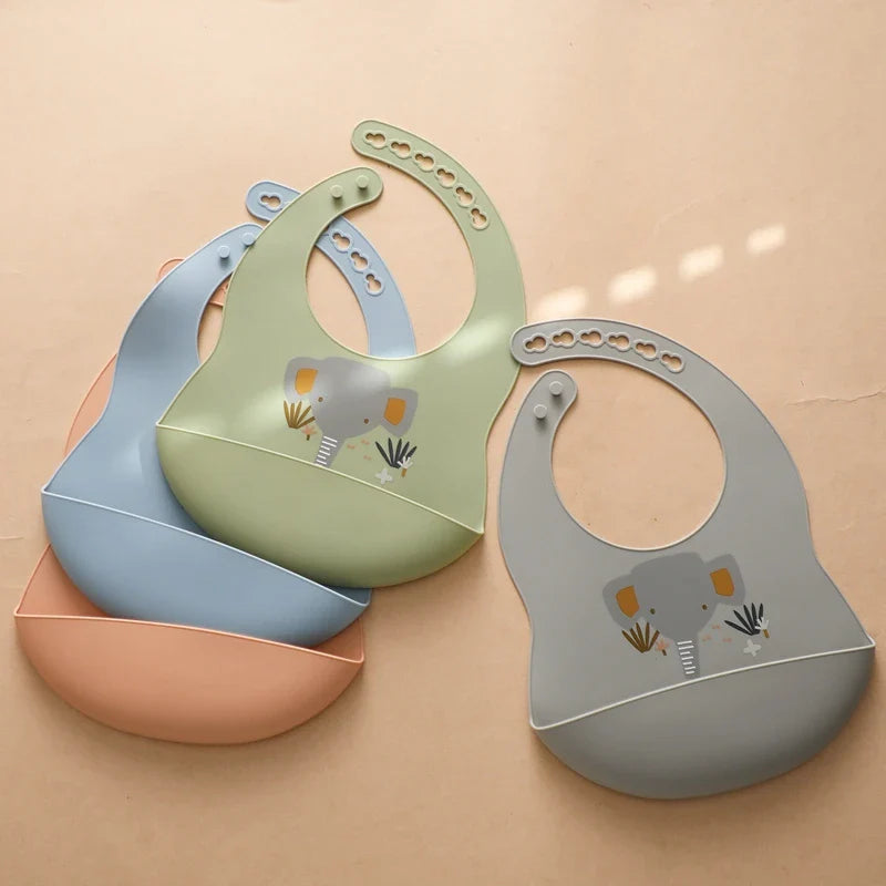 Drei Hello World Latz mit Motiv Silikon Grau [30x21cm], darunter Elefanten-Design und ein blauer Bib, liegen in Pastellfarben auf beigem Untergrund. Alle sind verstellbar und mit Knopfverschluss.