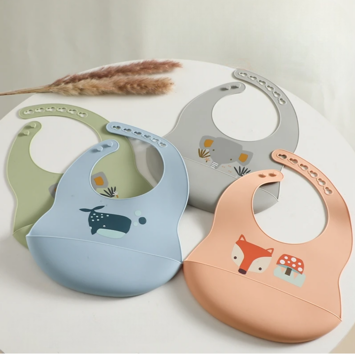 Der "Latz mit Motiv Silikon Orange" von Hello World (30x21cm) ist auf einer weißen Fläche abgebildet, die ein niedliches Naturmotiv zeigt und mit getrocknetem Pampasgras im Hintergrund akzentuiert ist.