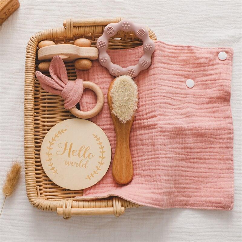 Das Lora Baby Geschenk zur Geburt von Hello World besteht aus einem Weidenkorb mit einem rosa Mulltuch, einer Babybürste aus Holz, einem Holz-Greifling-Beißring, einem Holzspielzeug und einer runden "Hello World"-Plakette - ideal für Neugeborene. Der Korb ist auf einer weißen, strukturierten Oberfläche abgebildet.