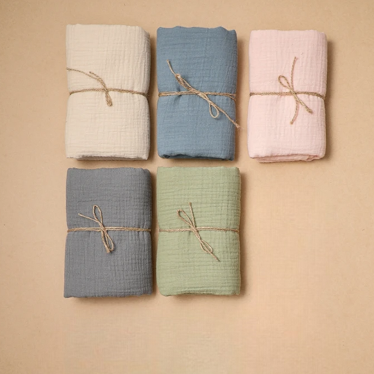 Fünf Hello World Multifunktionstücher Beige (55x65cm) in den Farben Creme, Blau, Rosa, Grau und Grün - perfekt als Spucktuch oder Stilltuch - sind mit Schnur gebunden und in zwei Reihen auf beigem Hintergrund angeordnet.