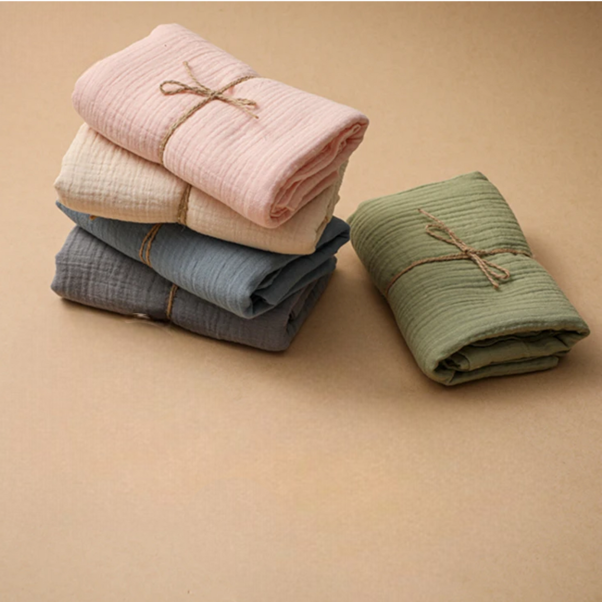 Das Hello World Multifunktionstuch Beige [55x65cm] liegt ordentlich gefaltet mit Bindfaden auf einer beigen Fläche, neben drei Musselin-Decken in Rosa, Weiß und Blau, die übereinander gestapelt sind - ideal als Still- oder Spucktuch.