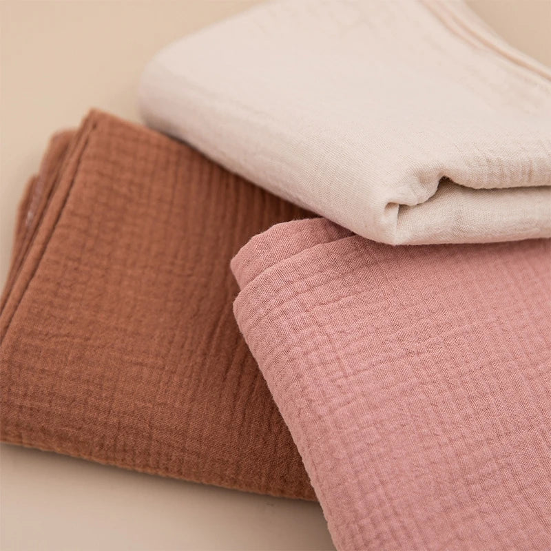 Drei fein säuberlich gefaltete Hello World Multifunktionstücher Braun [55x65cm] in hellem Creme, staubigem Rosa und warmem Braun sind auf einer neutralen Oberfläche gestapelt.