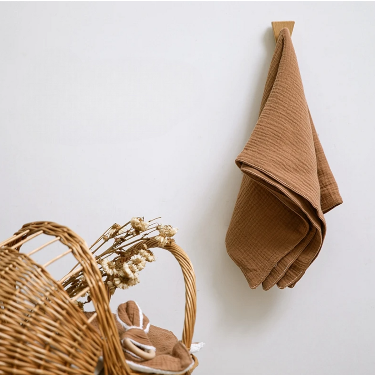 Ein Hello World Multifunktionstuch Braun [55x65cm] hängt an einem Wandhaken über einem Weidenkorb, der mit getrockneten Blumen und Stoffen gefüllt ist, vor einem schlichten weißen Hintergrund.