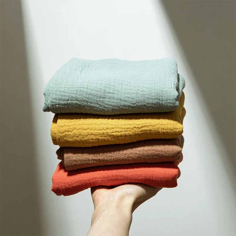 Eine Hand hält vier gefaltete, strukturierte Tücher - darunter das Hello World Multifunktionstuch Gelb [55x65cm] - in hellen Grün-, Gelb-, Braun- und Orangetönen vor einem sanft beleuchteten Hintergrund.