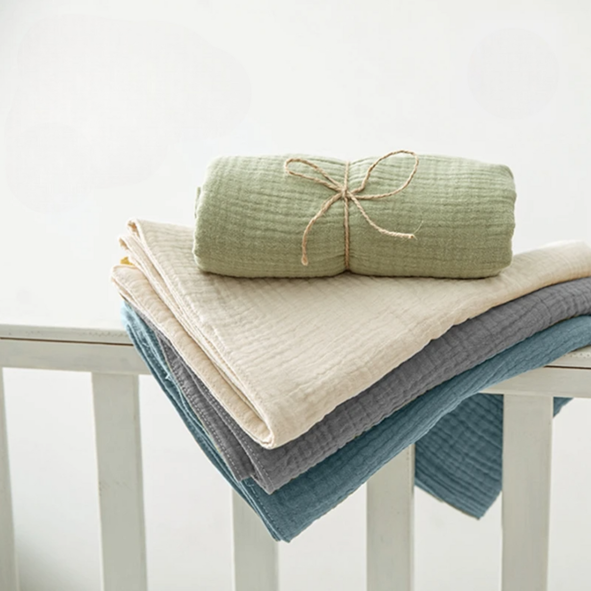 Ein Stapel von drei gefalteten Musselin-Decken in Creme, Grau und Blau liegt auf einem weißen Krippengitter, darüber das Hello World Multifunktionstuch Grün [55x65cm], ein umweltfreundliches Babytuch, das aufgerollt und mit einer Schnur zusammengebunden ist.