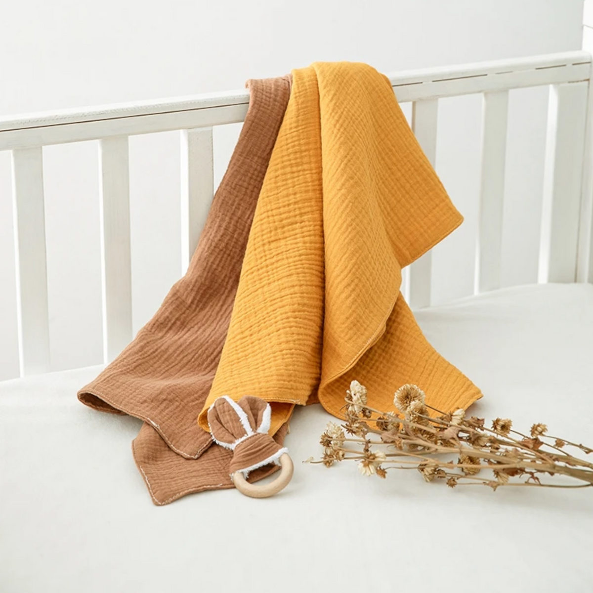 Zwei Musselin-Decken, eine senfgelb und eine braun, sind über einem weißen Kinderbett drapiert. Nebenan auf der Matratze liegt das Hello World Multifunktionstuch Orange [55x65cm], perfekt als Stilltuch, mit einem hölzernen Baby-Beißring und getrockneten Blumen.