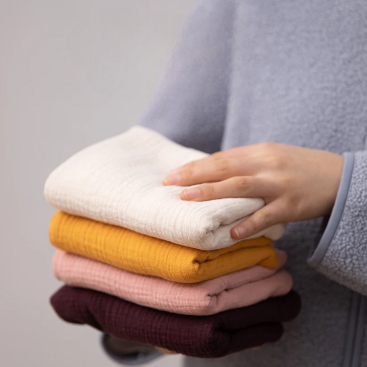 Eine Person in einem hellblauen Fleece hält einen ordentlich gefalteten Stapel Hello World Multifunktionstuch Orange [55x65cm] und drei weitere weiche Tücher in Creme, Gelb und Burgunderrot vor einem unifarbenen Hintergrund.