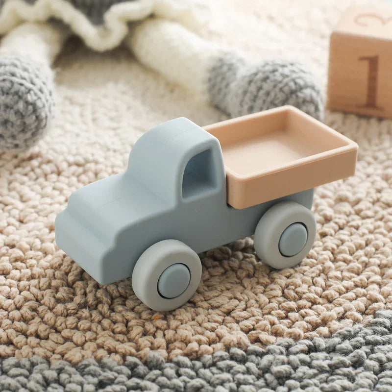 Ein kleines, pastellblaues Pickup Blau aus Silikon [11x5cm] von Hello World liegt auf einem beige- und cremefarbenen Teppich im Kinderzimmer, mit gestricktem Spielzeug und einem Holzklotz mit der Zahl 1 im Hintergrund.
