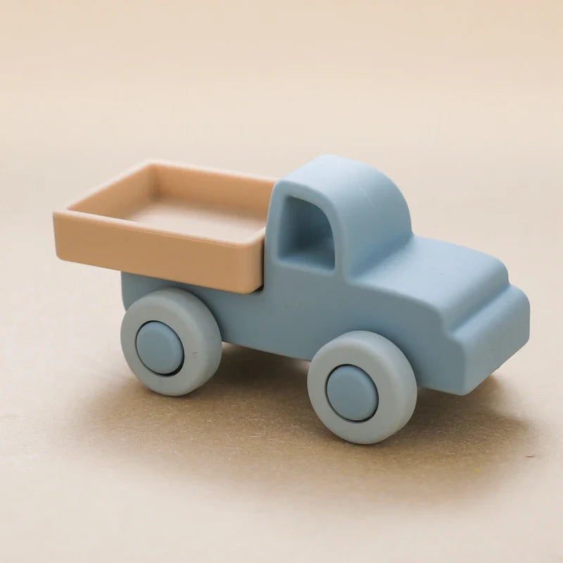 Der Hello World Pickup Blau aus Silikon [11x5cm] hat einen kleinen blauen Körper, eine beigefarbene Ladefläche und runde Räder - perfekt für jedes Kinderzimmer - auf einer neutralen, hellbraunen Oberfläche.