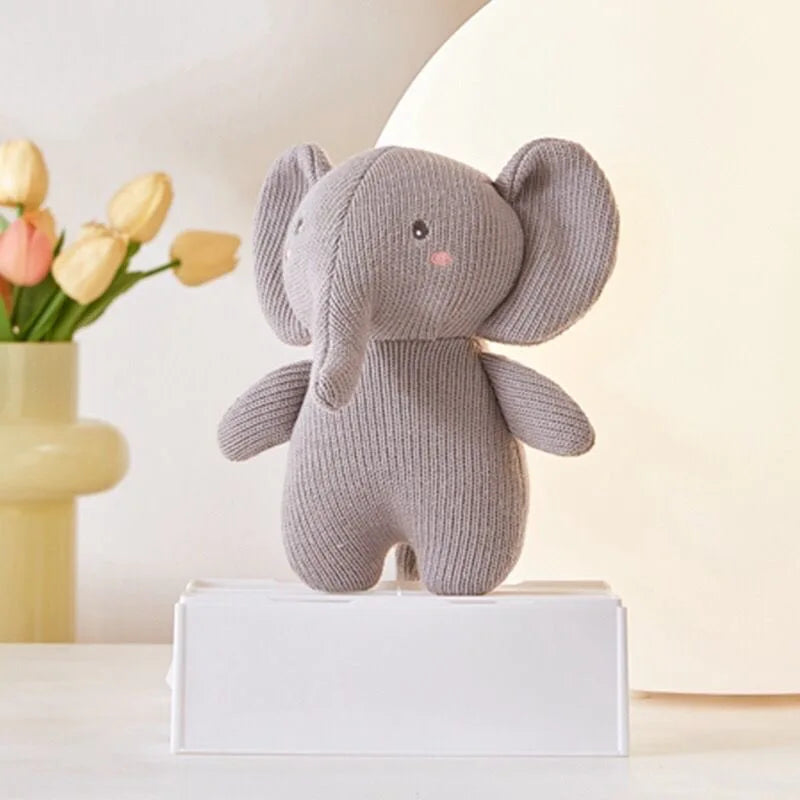 Der Plüschtier Elefant [14x16cm] von Hello World in Grau mit pinken Wangen steht auf einer weißen Box. Dahinter sind eine blassgelbe Vase mit Tulpen und ein sanft beleuchteter, cremeweißer Hintergrund zu sehen.
