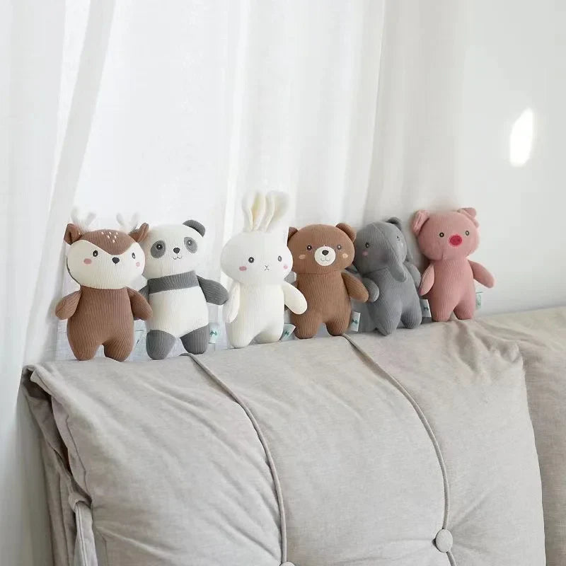 Sechs Plüschtiere - darunter das Hello World Plüschtier Schweinchen [18x14cm], ein Reh, ein Panda, ein Kaninchen, ein Bär und ein Elefant - sind auf einem hellgrauen Sofa mit weißen Vorhängen dahinter angeordnet.