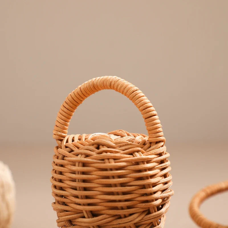 Ein kleiner geflochtener Weidenkorb mit einem geschwungenen Henkel ruht auf einem neutralen Hintergrund, dessen komplizierte Textur an die Handwerkskunst von Hello Worlds Rassel Eichel [6x13,5cm] erinnert, inspiriert vom traditionellen Naturinstrument Kinder.