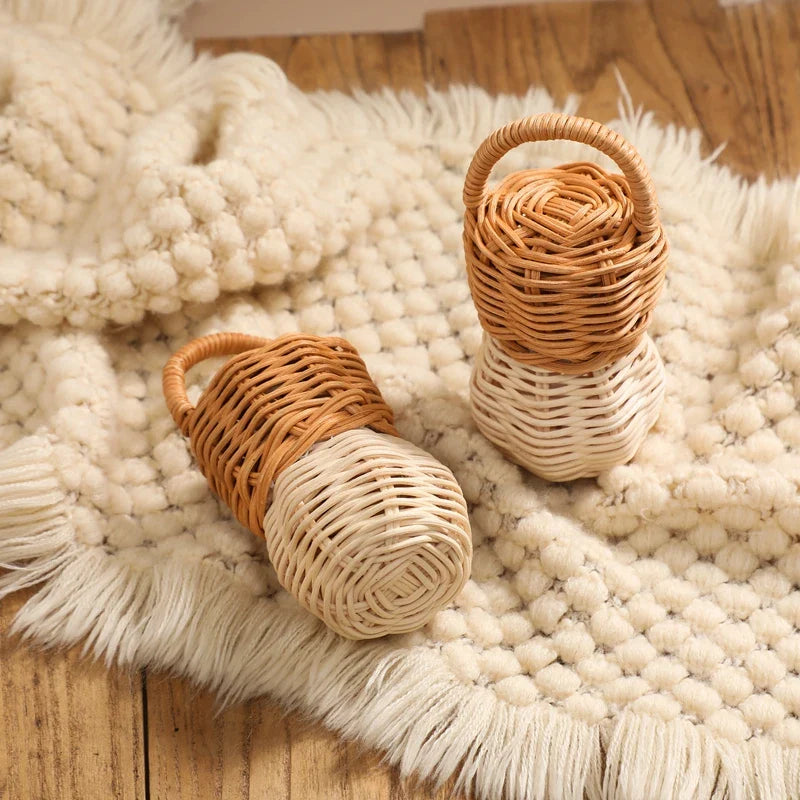 Zwei Hello World Rassel Eichel [6x13,5cm] aus geflochtenem Rattan, beide mit geschwungenen Griffen, ruhen auf einer cremefarbenen Fransendecke auf einer Holzfläche.