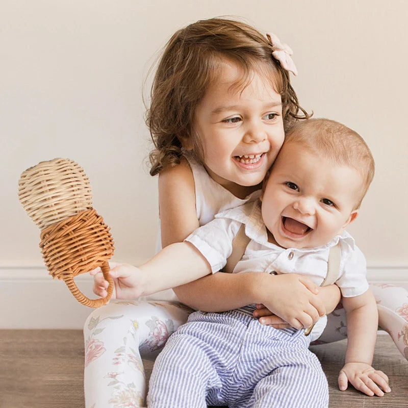 Ein lächelndes junges Mädchen umarmt ein lachendes Baby, das den Rassel Eichel [6x13,5cm] von Hello World hält. Sie sitzen auf dem Boden vor einer hellen Wand, beide sehen glücklich und verspielt aus.