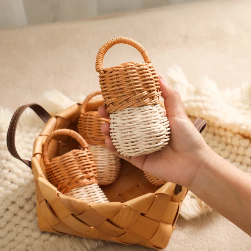 Eine Hand hält einen kleinen, zweifarbig geflochtenen Korb mit Henkel - ideal für die Aufbewahrung der Hello World Rassel Eichel [6x13,5cm]. Weitere Mini-Körbchen sind in einem größeren quadratischen Korb auf einem beigen, strukturierten Stoffhintergrund angeordnet.